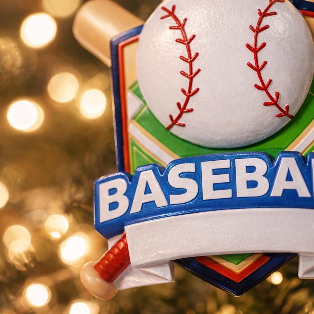 Personalized baseball Christmas ornament featuring a classic baseball and crossed bats design on a shield style background, customizable name area on the front banner and customizable year area on the baseball, glossy finish with raised 3D front details and flat 2D back, white hanging ribbon, displayed on a snowy baseball field themed holiday background with pine branches and festive decorations.