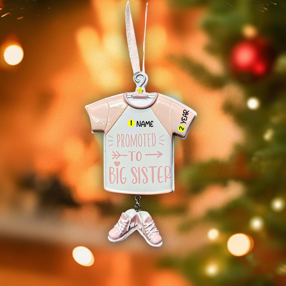 Decorative ornament shaped like a baby onesie with text, hanging against a Christmas tree background.