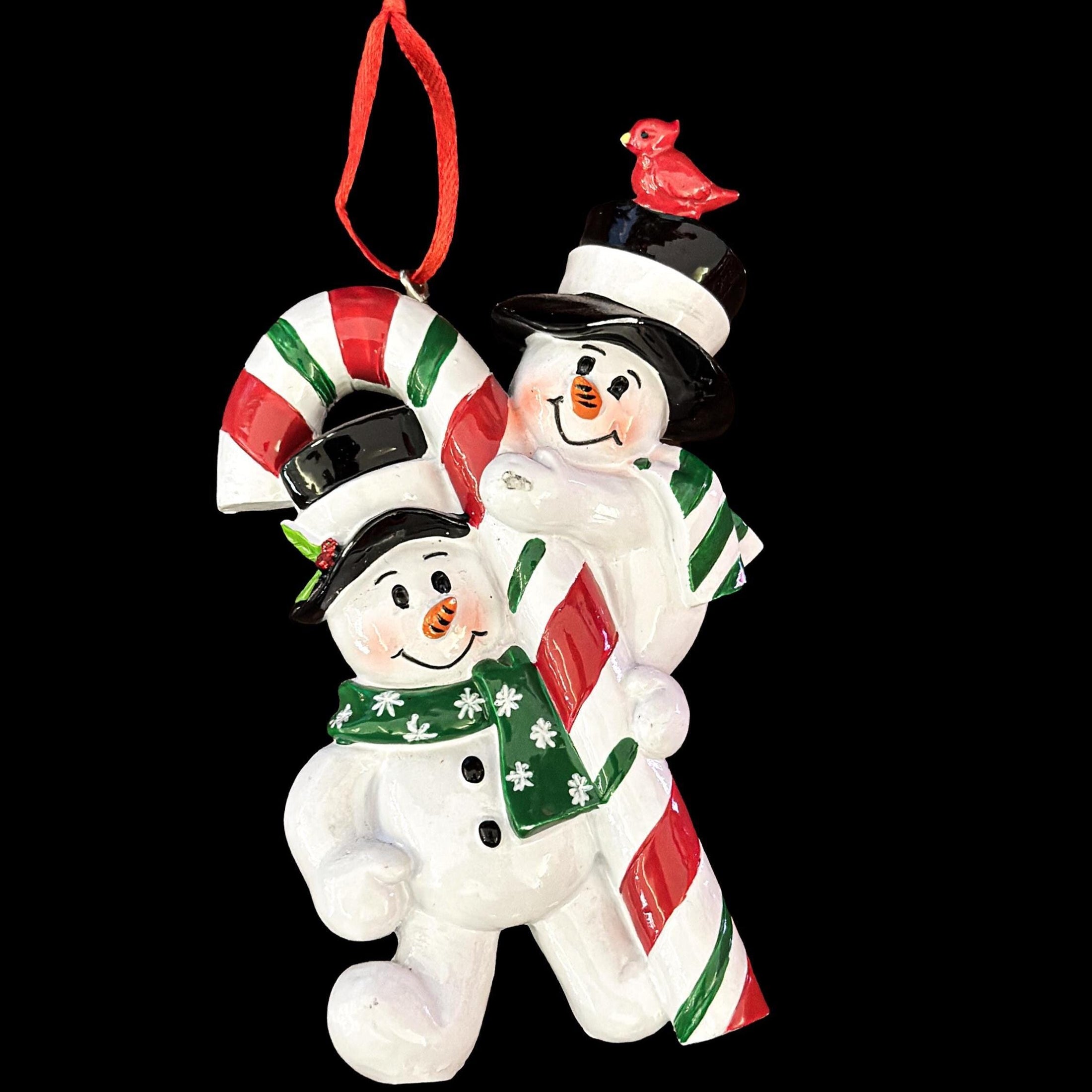 Christmas ornament featuring two snowmen holding a candy cane with a cardinal on top against a black background.