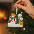Load image into Gallery viewer, Hand holding a Christmas ornament with snowmen and a tree against a blurred festive background.
