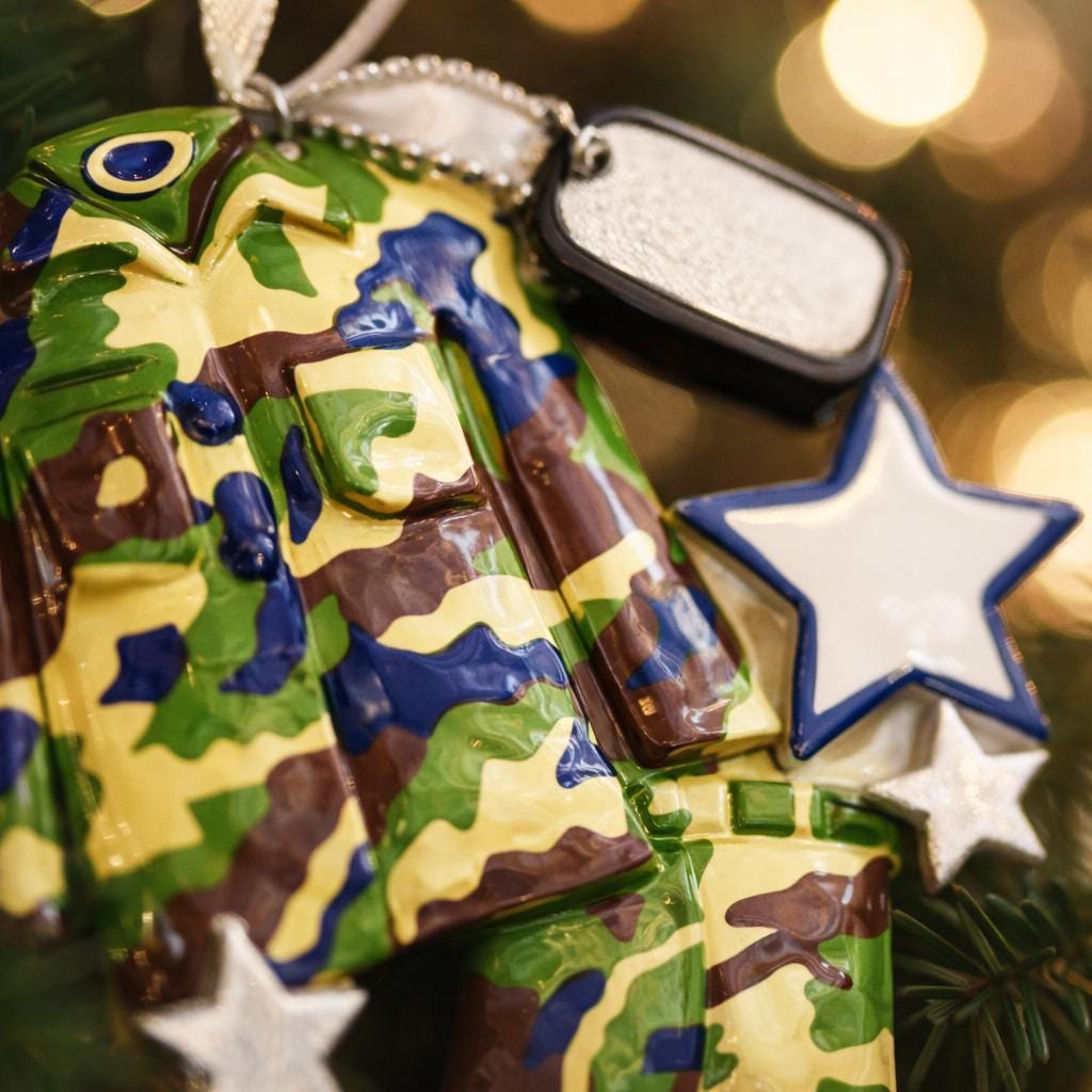 A glossy resin Army soldier uniform ornament in green brown and blue camouflage colors features a military jacket and pants shape with raised buttons and pockets, white and blue star accents, light snow texture, a smooth painted cartoon style finish, and handwritten personalization showing a name and year on the attached tag, designed as a mostly 2D ornament with raised 3D details on the front only for Christmas tree decor.