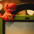 Load image into Gallery viewer, Red dinosaur ornament holding a yellow object on top of a green electronic device.
