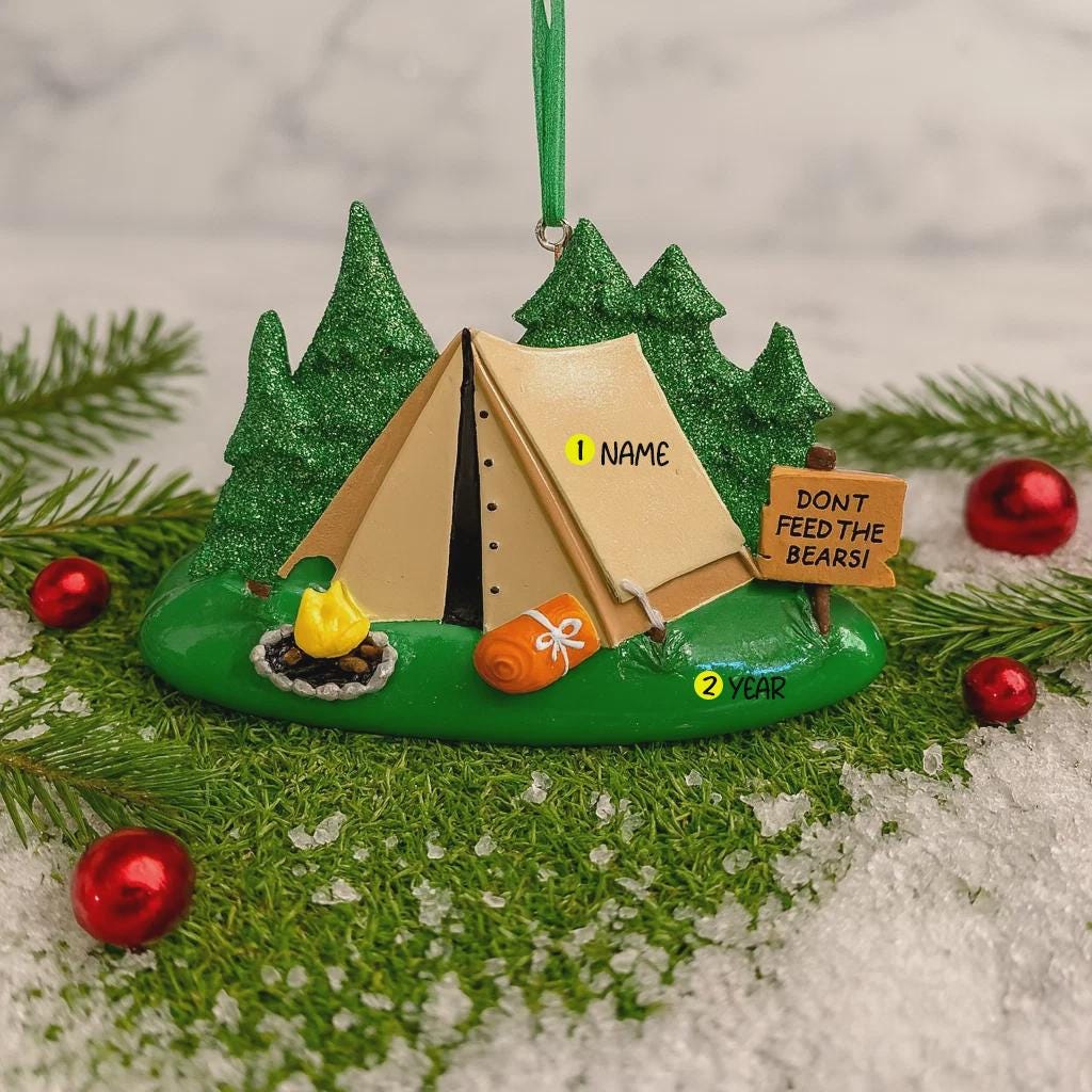 Camping-themed ornament with a tent, fire, and sign on a green base with trees and red ornaments.