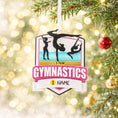 Load image into Gallery viewer, Personalized gymnastics Christmas ornament featuring elegant gymnast silhouette figures in motion, customizable name area on the front banner and customizable year area above the banner, glossy finish with raised 3D front details and flat 2D back, white hanging ribbon, displayed on a soft pink snowy holiday background with festive ornaments greenery and warm Christmas decor.

