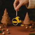 Load image into Gallery viewer, Yellow and black tape measure ornament held by a hand against a Christmas-themed background.

