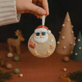 Load image into Gallery viewer, Hand holding a decorative ornament with a Santa Claus face on a brown surface with Christmas-themed decorations.
