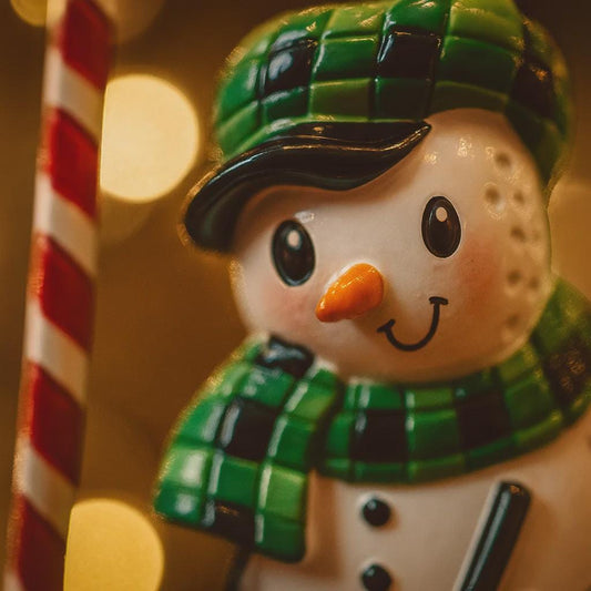 Decorative snowman ornament with green hat and scarf, standing next to a candy cane.