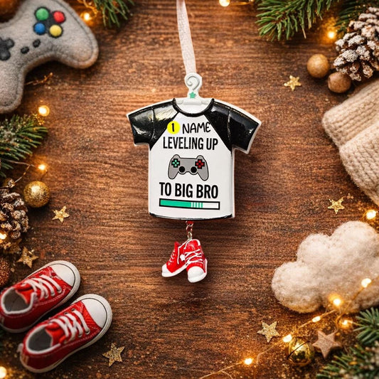 Decorative ornament shaped like a t-shirt with text, surrounded by Christmas decorations on a wooden surface.