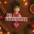 Load image into Gallery viewer, Personalized Babysitter Christmas ornament featuring a detailed raised 3D front design with glitter pink lettering and a charming girl figure. Custom name is added on the shirt and year is written on the hanging heart charm. Bright pink and white holiday colors stand out beautifully on the tree. Flat 2D back for easy hanging. Perfect keepsake gift for babysitter appreciation, nanny thank you present, childcare provider Christmas decor.
