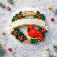 Load image into Gallery viewer, Personalized cardinal wreath ornament featuring a vibrant red cardinal bird surrounded by green holly leaves and red berries. Designed with a smooth glossy finish and a raised 3D front with a flat back for easy hanging. Includes free handwritten personalization with one name and year displayed on the banner at the top. Styled in a warm festive holiday setting with soft Christmas lights candy details and seasonal decor. A meaningful Christmas tree ornament symbolizing remembrance love and connection perfect 
