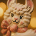 Load image into Gallery viewer, A glossy resin baby girl llama ornament features a white smiling llama with soft pink accents, raised textured fur details, pink bow and scarf elements, glitter highlights, and a smooth painted cartoon style finish, styled with warm holiday lights and nursery themed props, handwritten personalization showing the baby name on the front banner and the year placed on the llama body, designed as a mostly 2D ornament with raised 3D details on the front only.
