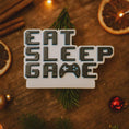 Load image into Gallery viewer, Personalized Eat Sleep Game Christmas ornament with a raised 3D front featuring bold block-style lettering and a detailed game controller icon. The front design is fully three dimensional, creating depth and texture, while the back is flat 2D for easy hanging and durability. White base with black raised letters and subtle colored accents. Includes hand-added personalization with name and year or family name. Gaming-themed holiday ornament displayed on snowy festive background with warm lights, designed for 

