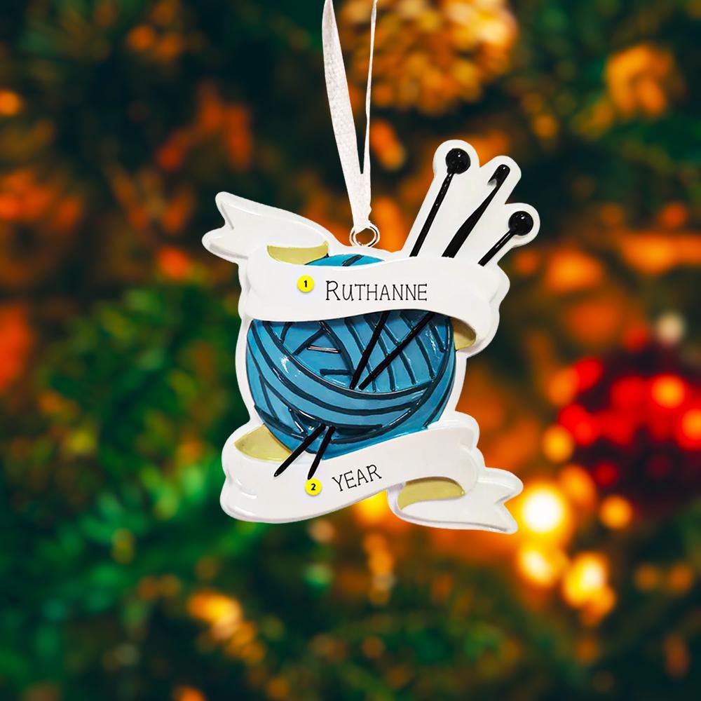 A cozy knitting yarn ornament featuring a detailed blue ball of yarn wrapped with flowing ribbon and raised knitting needles. The ornament has a sculpted 3D front with realistic yarn texture and smooth glossy finish, while the back is flat and 2D for easy hanging. Personalized with a handwritten name and year, this resin ornament is displayed in a warm festive setting with soft holiday lights, creating a heartfelt and nostalgic feel. Perfect for grandmother gifts, knitting lovers, and handmade craft themed 
