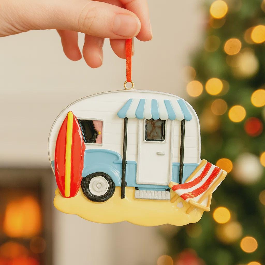 Decorative ornament shaped like a camper with a surfboard and beach chair, held against a blurred Christmas tree background.