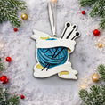 Load image into Gallery viewer, A cozy knitting yarn ornament featuring a detailed blue ball of yarn wrapped with flowing ribbon and raised knitting needles. The ornament has a sculpted 3D front with realistic yarn texture and smooth glossy finish, while the back is flat and 2D for easy hanging. Personalized with a handwritten name and year, this resin ornament is displayed in a warm festive setting with soft holiday lights, creating a heartfelt and nostalgic feel. Perfect for grandmother gifts, knitting lovers, and handmade craft themed 
