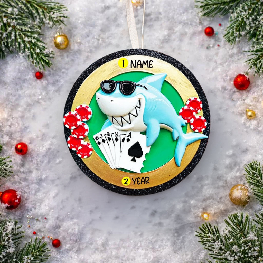 Personalized poker ornament featuring a cool shark character wearing sunglasses with playing cards and red casino chips. Designed in a round ornament style with a glossy finish. Includes free handwritten personalization with one name and year displayed on the front. Shown hanging with a ribbon in a festive holiday setting with greenery snow and seasonal decor. 3D raised front and flat back design perfect for Christmas trees and gifts for poker players casino lovers and card game fans.