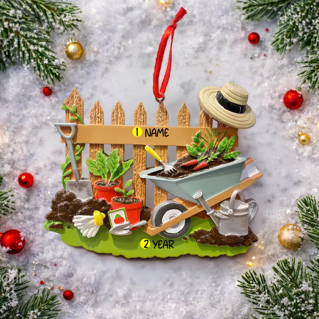 Personalized gardener ornament featuring a detailed garden scene with wooden fence wheelbarrow potted plants gardening tools and soil elements. The design includes a customizable name on the fence and a year on the base making it a thoughtful keepsake for garden lovers. The ornament has a warm rustic farmhouse style and is displayed with a red hanging ribbon suitable for Christmas trees seasonal decor and gift displays.
