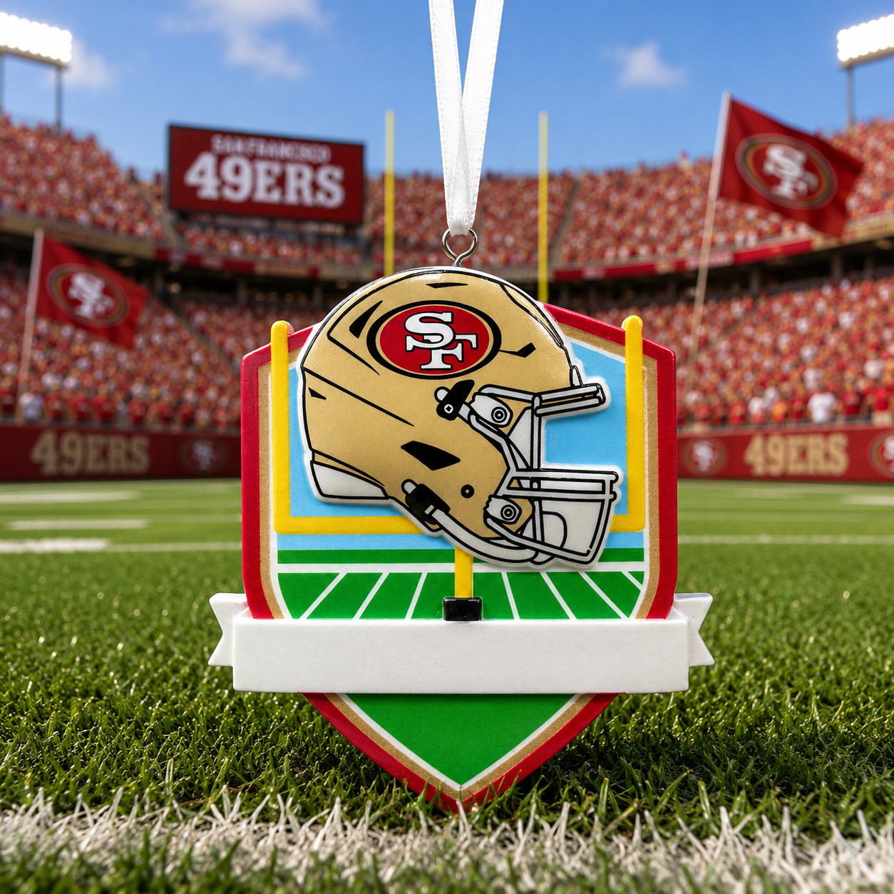 A San Francisco 49ers themed ornament shaped like a football helmet mounted on a shield over a football field, hanging by a white ribbon in front of a stadium background.