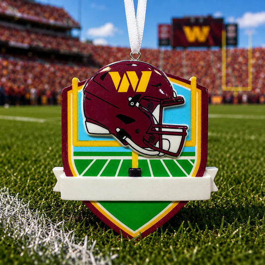 A football-themed ornament shaped like a helmet and shield, hanging on a grassy field with a blurred stadium background. The ornament features a burgundy helmet with a yellow 'W' logo, green field details, and a white hanging ribbon.