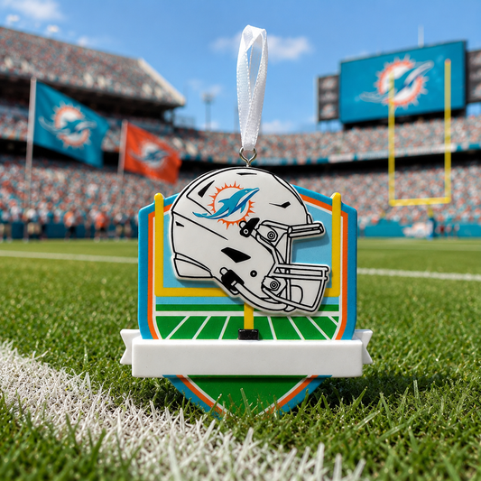 A Miami Dolphins themed football ornament displayed on a green turf field with stadium stands in the background. The ornament features a white football helmet with the team's blue and orange logo, set against a graphic of a football field and uprights.